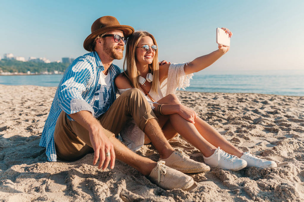 young attractive smiling happy man and woman in sunglasses sitting on sand beach taking selfie photo on phone camera, romantic couple by the sea on sunset, boho hipster style outfit - Fotografie, Obrázek