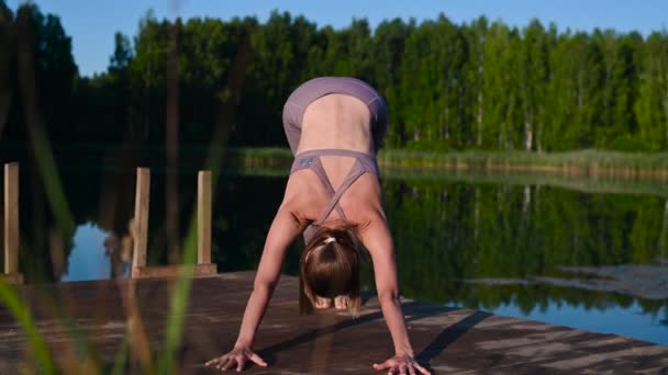 Młoda kobieta nad jeziorem ćwicząca jogę porusza się na drewnianej platformie. Ładna młoda kobieta ćwicząca w naturze, zdrowy styl życia młodzi ludzie pozytywne wibracje. - Materiał filmowy, wideo