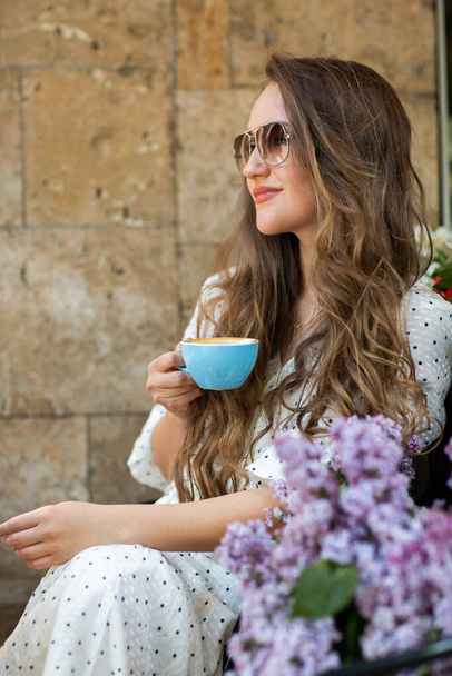 Beautiful Woman in sunglasses Enjoying the morning coffee near lilac blossom. Cute Model and Flowers. Aromatherapy and Spring Concept. Girl drink coffee at coffee shop. - Foto, imagen