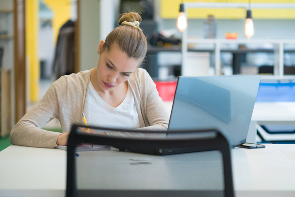 Mujer joven atractiva usando el ordenador portátil mientras está sentada en su escritorio. Joven mujer de negocios americana sentada en la oficina y trabajando en el portátil. - Foto, Imagen