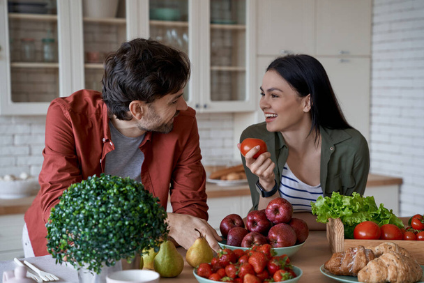 Une saine alimentation. Jeune belle femme tenant tomate fraîche et regardant son mari tout en préparant des aliments sains ensemble dans la cuisine moderne à la maison - Photo, image
