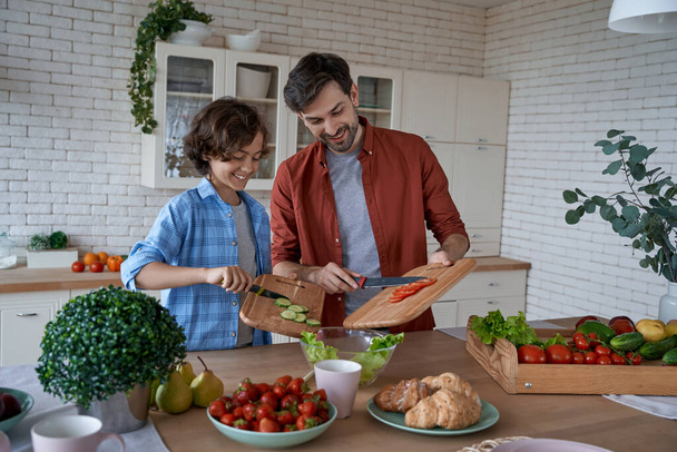 Kochen mit Papa. Junger Vater und sein glücklicher Sohn bereiten einen Salat zu, während sie zu Hause in der modernen Küche stehen und Zeit miteinander verbringen - Foto, Bild