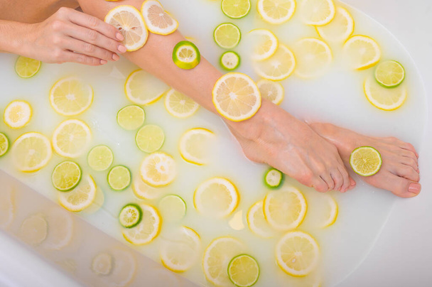 Unrecognizable woman takes a milk bath with lemons and limes. Citrus spa. Body care. Skin whitening. Female legs top view. - Fotografie, Obrázek
