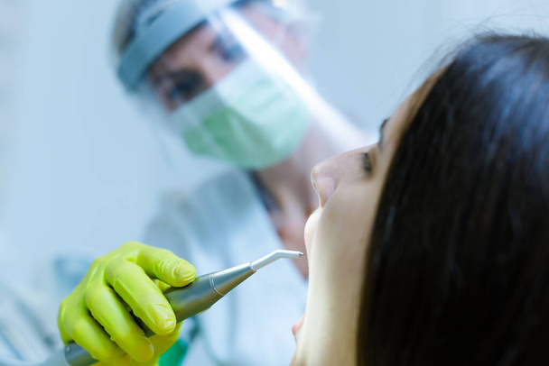 Vrouwelijke tandarts draagt gezichtsmasker, handschoenen en vizier als coronavirus veiligheidsmaatregel controle tandheelkundige gezondheid van jonge vrouw - Foto, afbeelding