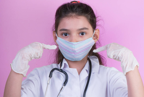 Μικρό αστείο κορίτσι με ένα κολλαριστό στηθοσκόπιο ενώ φορούσε τη στολή του γιατρού. πολύχρωμη μάσκα, πανόραμα. Copyspace. πορτρέτο λίγο γιατρό. covid19. γάντια που μπορούν να αφαιρεθούν  - Φωτογραφία, εικόνα