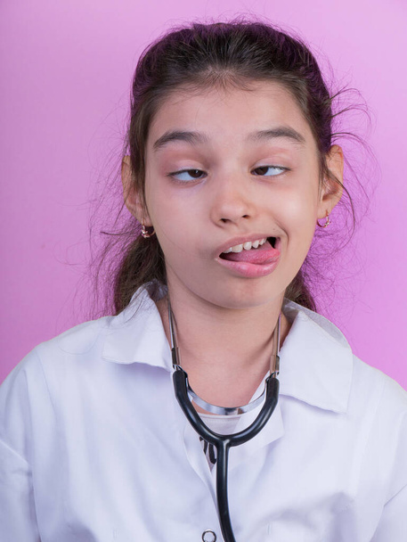 Little funny girl with a collorful stethoscope while wearing Doctor's uniform. Excitement and fascination concept. Coronavirus background. foolish grimaces comical crazy gestur.  Funny expression.  - Photo, Image