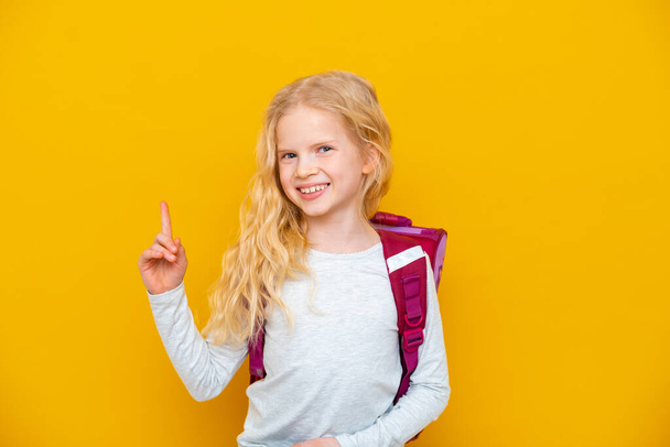 Back to school. Portrait of blonde school girl with bag and books. Pointing finger up. Yellow studio background. Education. smiling at camera. - Фото, изображение