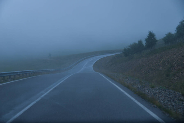 the roadbed is uneven, winding, passes through mountains and cliffs. Clouds are dark, low, with rain. A thick white fog is spreading along the road. - Fotoğraf, Görsel