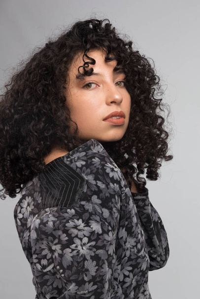 young latin woman with curly hair and brown skin from side, wearing black patterned clothing, studio with gray background - Photo, Image