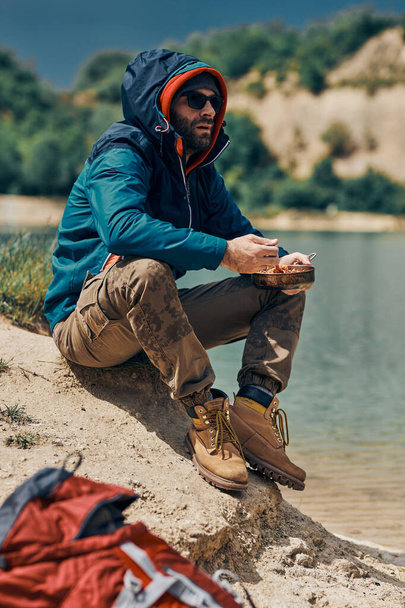 キャンプ旅行で湖のそばに座って昼食をとる若者. - 写真・画像