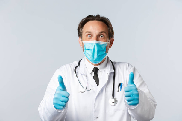 Covid-19, preventing virus, healthcare workers and vaccination concept. Excited and surprised happy doctor in medical mask and gloves, showing thumbs-up thrilled, hear great news, agree and approve - Fotoğraf, Görsel
