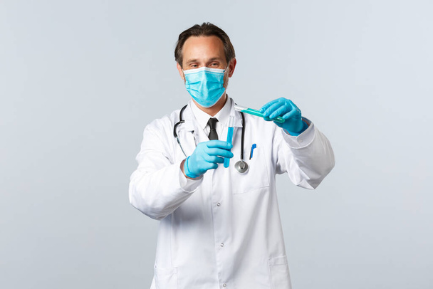 Covid-19, Prävention von Viren, Gesundheitspersonal und Impfkonzept. Lächelnder Arzt in medizinischer Maske und Handschuhen, der chemische Zutaten in ein anderes Reagenzglas gießt und Impfstofftests durchführt - Foto, Bild