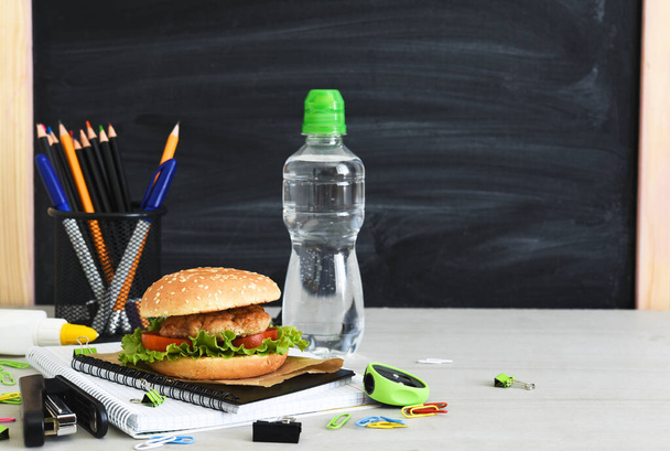 Terug naar school. Quarantaineonderwijs. lunchbox met hamburger, water en schoolaccessoires op de achtergrond van het schoolbestuur. - Foto, afbeelding