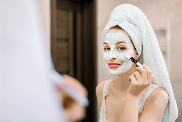 Beauty skin care, cosmetics and hygiene concept. Young woman with towel on head, looking in the mirror, holding cosmetic brush and applying facial clay mud mask to her face in bathroom - Foto, Imagem