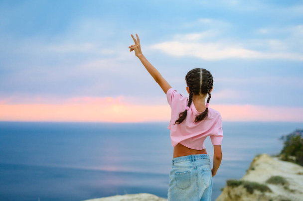 A girl in a pink T-shirt stands with her hand held high on the edge of a mountain. Happy little girl looking at the sunset and the sea - Photo, Image