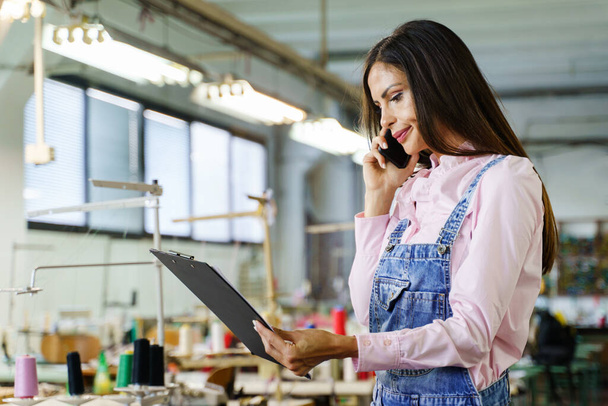 Young female entrepreneur analyze data working at sewing factory - Side view on woman holding files clip documents while making a phone call at work - Copy space business production planning concept - Фото, зображення