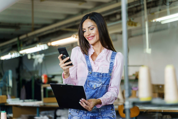 Happy young entrepreneur smiling at work while holding mobile phone - Female manager or worker making video call or texting in factory - success achievement happiness and independence equality concept - Photo, image