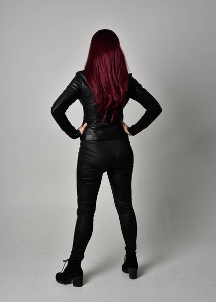 Full length portrait of  girl with red hair wearing black leather jacket, pants and boots. Standing pose facing away from the camera, isolated against a grey studio background. - Foto, Bild