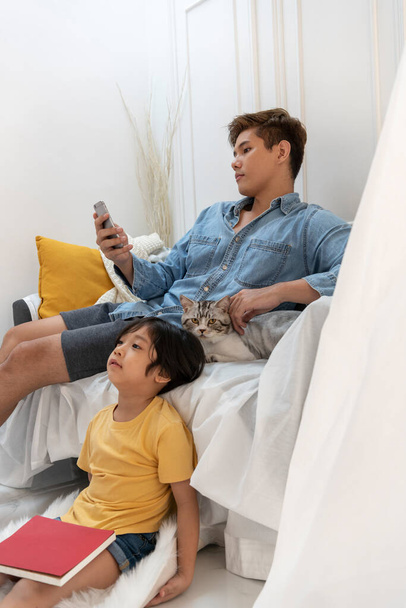 Boldog ázsiai család macskával. Apa okostelefont használ, miközben egy macskához dörgölőzik. Fiam belefáradt egy könyv olvasásába, a TV-be néz. - Fotó, kép