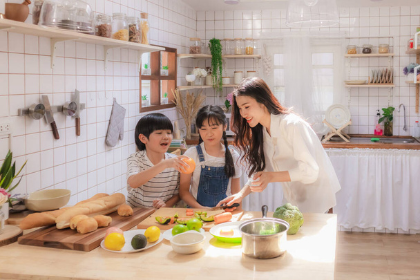 Счастливая и милая азиатская семья готовит еду на кухне дома вместе. Улыбающиеся мать, дочь и сын наслаждаются приготовлением пищи. Связь любви между родителями и детьми. - Фото, изображение