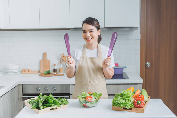 Усміхнена молода жінка в фартусі стоїть на сучасній кухні готуючи салат
 - Фото, зображення