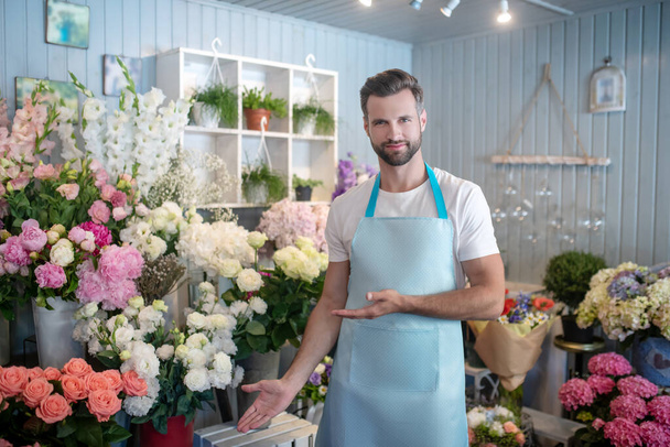 Bearded florist in apron standing in front of flower stand showing flowers - Fotoğraf, Görsel