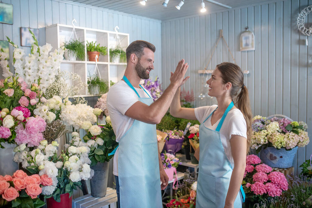Bearded male and brown-haired female standing intside flower shop giving high five - Фото, изображение