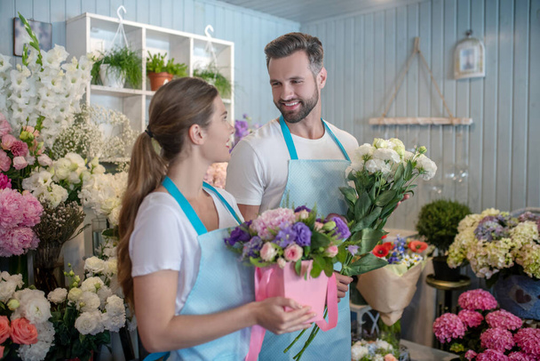 Bearded male and brown-haired female in aprons holding flowers, looking at each other, smiling - Foto, Imagem