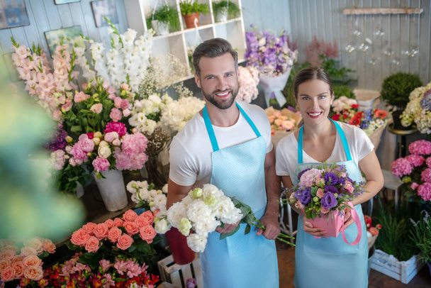 Bearded male and brown-haired female in aprons, holding bouquet and box of flowers, smiling - Фото, изображение
