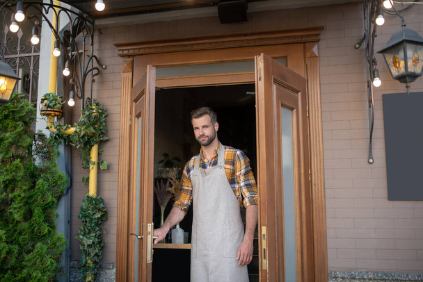 Young bearded male in grey apron holding handle, opening wooden door - Fotoğraf, Görsel