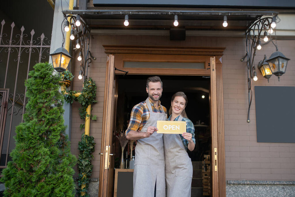 Young waitress and bearded waiter in grey aprons standing outside, holding yellow open sign - Foto, Bild