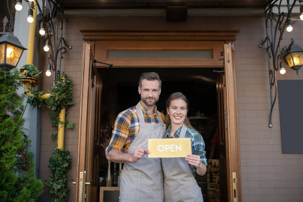 Young waitress and bearded waiter standing outside, holding open sign, welcoming clients - Foto, afbeelding