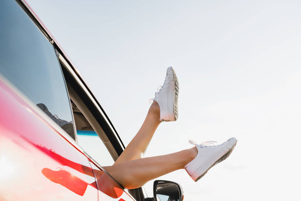 Playful girl sticking out feet in white sneakers from vehicle at sky background, happy traveling adventure. - Zdjęcie, obraz