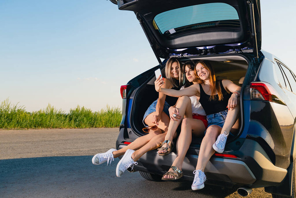Three girls take a selfie together. Blonde, brunette and red haired young women sit down and relax at vehicle back, talking during road adventure trip in nature. - Photo, Image
