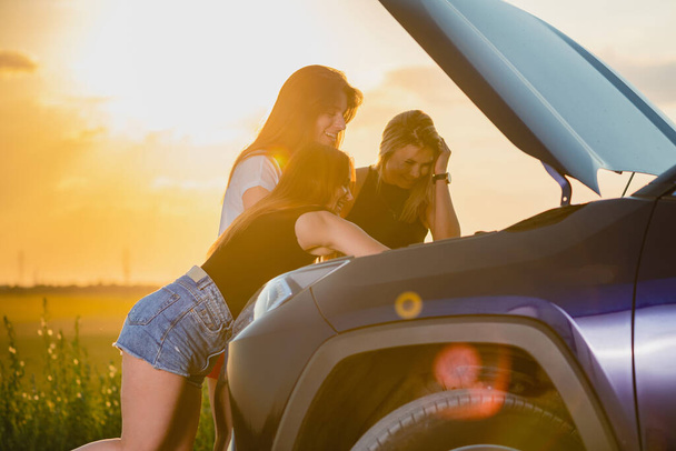 Ženské kamarádky uvízly v terénu během výletu, kontrolu a snaží se opravit vozidlo s otevřenou kapotou při západu slunce světlo, cestovní dobrodružství - Fotografie, Obrázek
