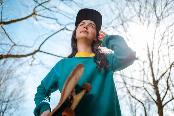 Een jonge blanke hippe vrouw poseert met een skateboard in haar handen en kijkt in de verte. Op de achtergrond, blauwe lucht, bomen en zon. Onderaanzicht. Concept van sportieve levensstijl en straatcultuur. - Foto, afbeelding