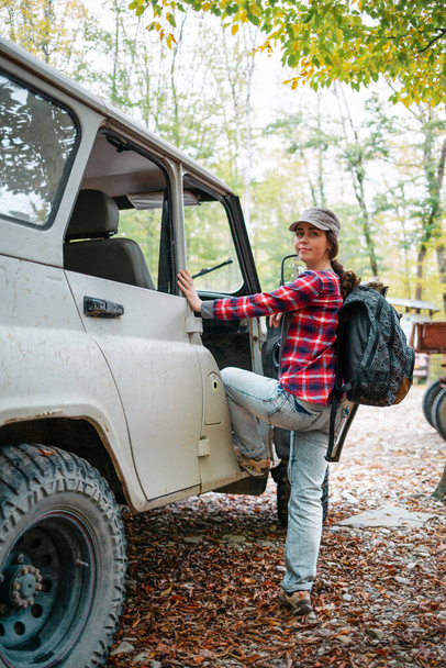 Una giovane donna caucasica con uno zaino sale su una jeep. Alberi sullo sfondo. Vista laterale. Verticale. Concetto di viaggio in auto e turismo. - Foto, immagini