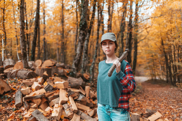 Preparation for the heating season. A young woman stands with an axe on her shoulder. In the background, a pile of firewood and autumn forest. - Photo, image