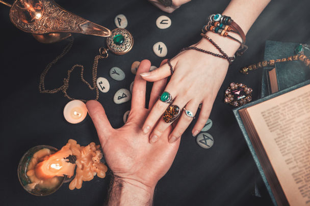 Astrologie und Esoterik. Eine Wahrsagerin liest einem Mann die Hand. Auf schwarzem Hintergrund liegen Runen, eine Kerze, Amulette und ein Buch. Der Blick von oben. - Foto, Bild