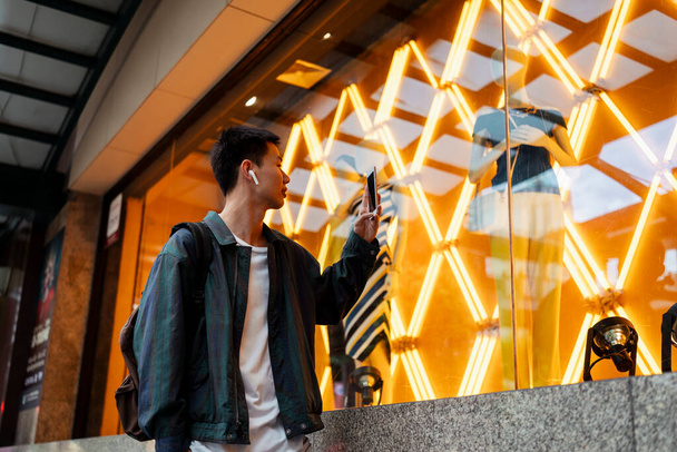 Homme aux cheveux courts utiliser smartphone pour prendre une photo de la boutique de robe avec lumière au néon orange. - Photo, image