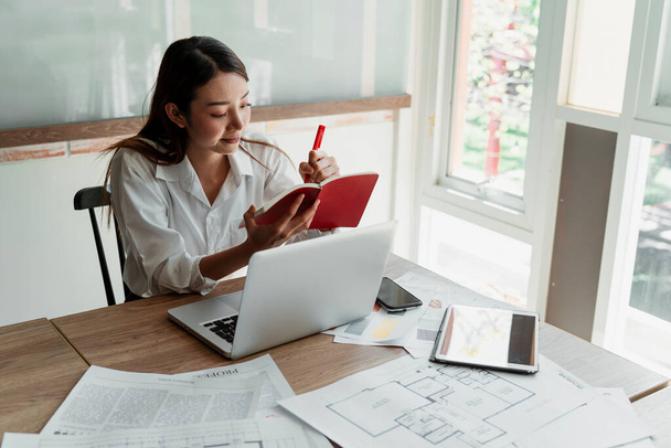 Długie włosy projektantka kobieta w białej koszuli siedzi w biurze wziąć notatkę do czerwonego notatnika podczas gdy ona dostaje świetny pomysł. - Zdjęcie, obraz