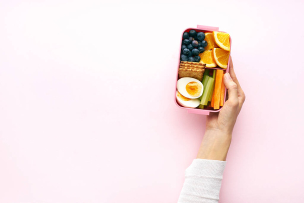 Female hands holding lunchbox with egg and orange fruits with blueberries and crackers , top view - Foto, afbeelding