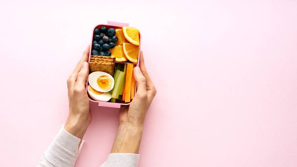 Female hands holding lunchbox with egg and orange fruits with blueberries and crackers , top view - Фото, зображення