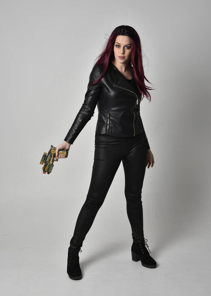 Ganzkörperporträt eines Mädchens mit roten Haaren, das schwarze Lederjacke, Hose und Stiefel trägt. Stehende Pose mit einer Waffe, isoliert vor grauem Studiohintergrund. - Foto, Bild