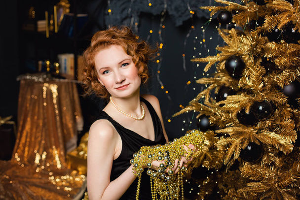 Model girl holding beads garlands on the background of a Christmas tree with gold decorations. Stylish women's, fashionable evening wear and accessories, makeup. Happy New Year. - Fotografie, Obrázek