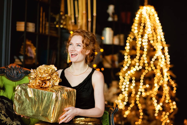 Egy boldog lány karácsonyi ajándékdobozokkal ajándékokat tart a háttérben egy karácsonyfa arany dekoráció. Stílusos női, divatos estélyi viselet és kiegészítők, smink. Boldog új évet!. - Fotó, kép