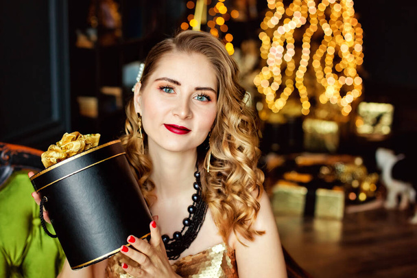 Model dziewczyna z pudełkami prezentów świątecznych trzymając prezenty na tle choinki ze złotymi dekoracjami. Stylowe damskie, modne stroje wieczorowe i akcesoria, makijaż. Szczęśliwego Nowego Roku. - Zdjęcie, obraz
