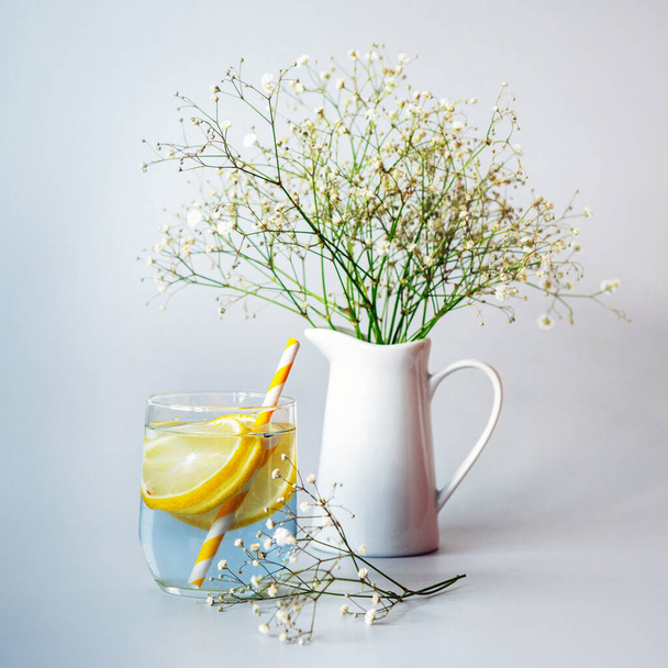 Ένα ποτήρι λεμονάδα με καλαμάκι και λουλούδια Gypsophila σε λευκή κανάτα - Φωτογραφία, εικόνα
