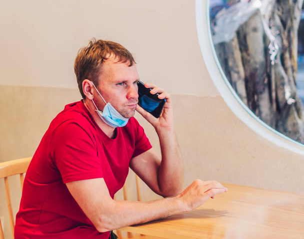 Young serious pensive attentive mean in medical face mask tense thinking speak answer phone in red t-shirt in cafe. Real people expression, life problems solving, communication during Covid Pandemic - Zdjęcie, obraz
