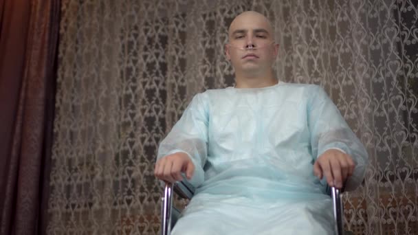 Un joven calvo con cáncer mira a la cámara y reza. El paciente dobló los brazos y oró en silla de ruedas en su casa. Pérdida de cabello debido a quimioterapia. - Metraje, vídeo